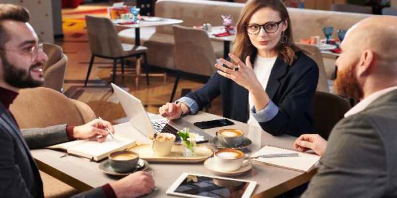 Group of business colleagues sitting at table with coffee cups in cafe and discussing business ideas using portable devices
