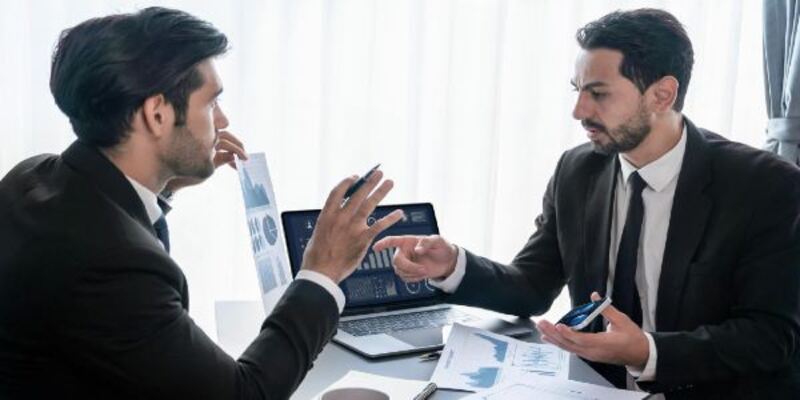 Two men in suits are sitting at a desk, one of them is holding charts