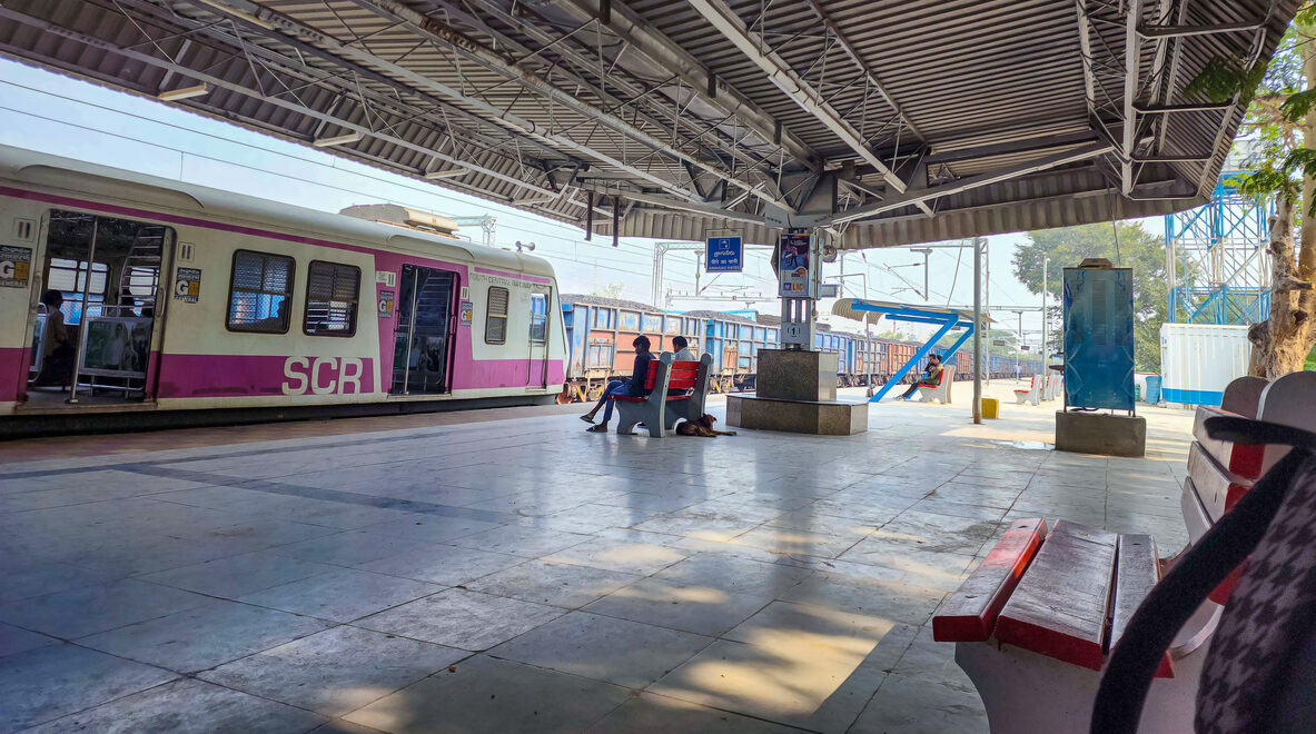 Two people sat on the wooden bench under the gray shelter, patiently awaiting the arrival of the train.In the background, a local train arrives at the station at Hyderabad.,India. Track doubling projects for AP and Telangana