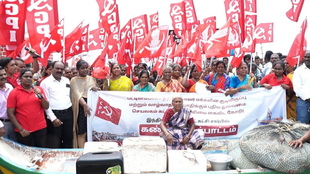 A large group of fishermen and supporters protest while holding red CPI flags and a Tamil banner, with fishing nets and boats in the foreground.