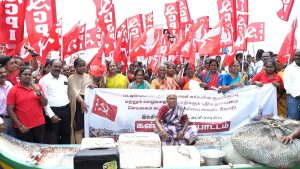 A large group of fishermen and supporters protest while holding red CPI flags and a Tamil banner, with fishing nets and boats in the foreground.