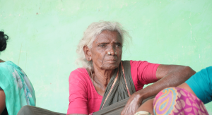 Portrait of Perumayi, an elderly woman from Thandiyakulam, seated against a green wall.