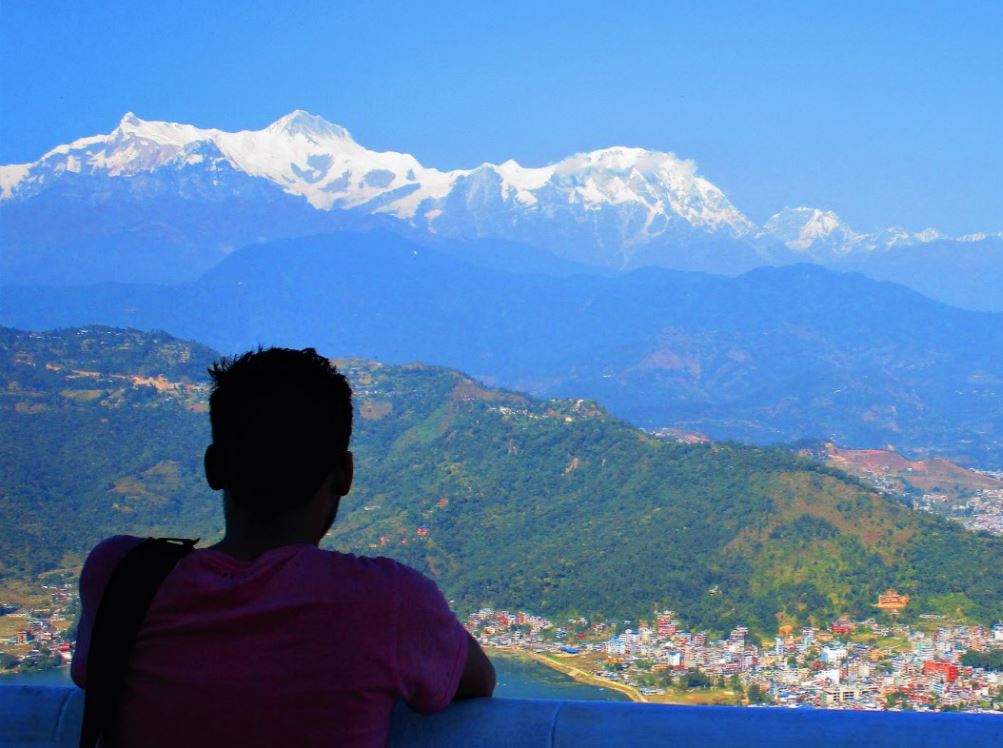 Man standing at edge looking at the snow covered mountains peak of himalayan region with local village at the foot of green mountain